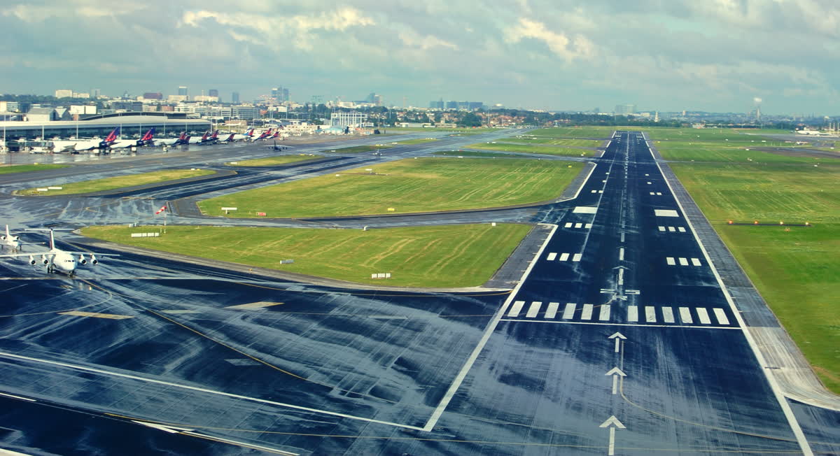 Yağmur sonrası ıslak bir havalimanı pisti, yeşil çim alanlar ve uzakta terminal önünde park etmiş yolcu uçakları